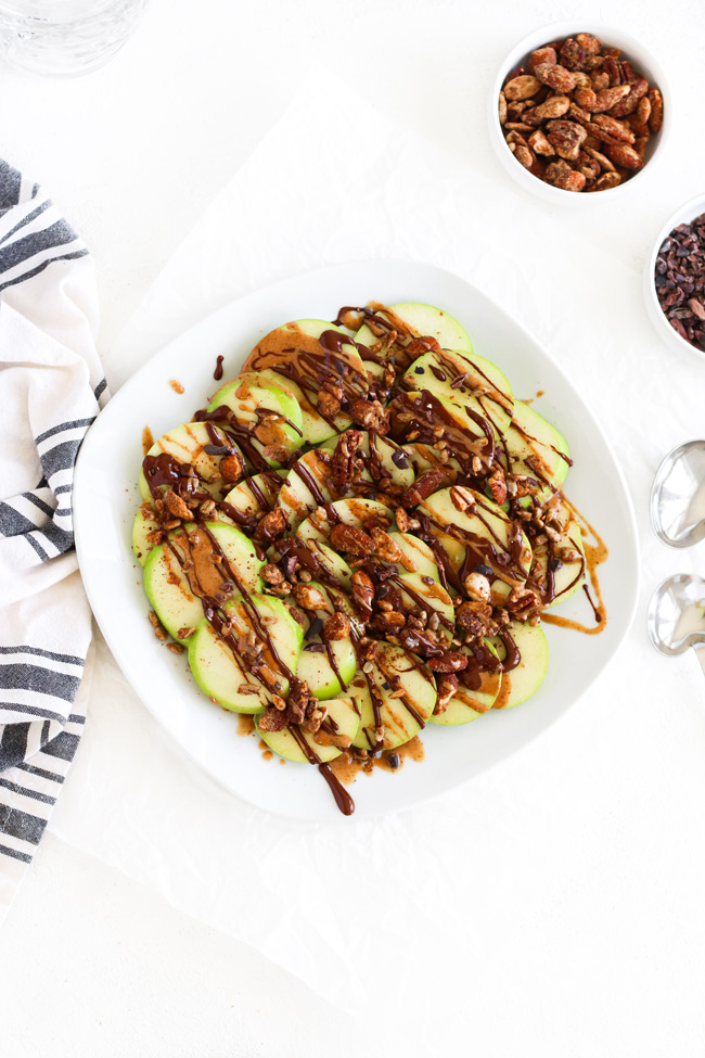 Green Apple Breakfast Nachos topped with granola on a white plate next to a black and ivory striped napkin and dishes of granola and carob nibs.