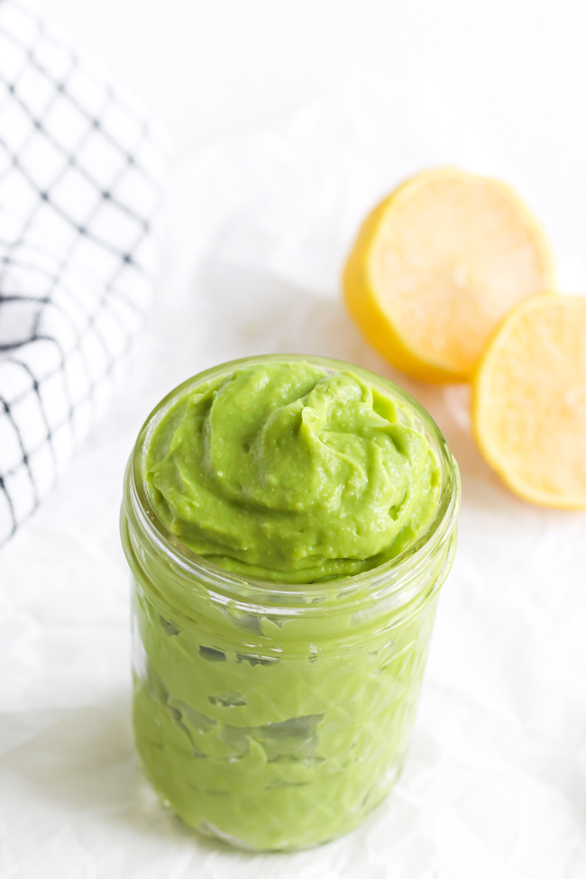 Avocado Mayo in a small glass jar next to fresh lemons and a black and white plaid cloth napkin.