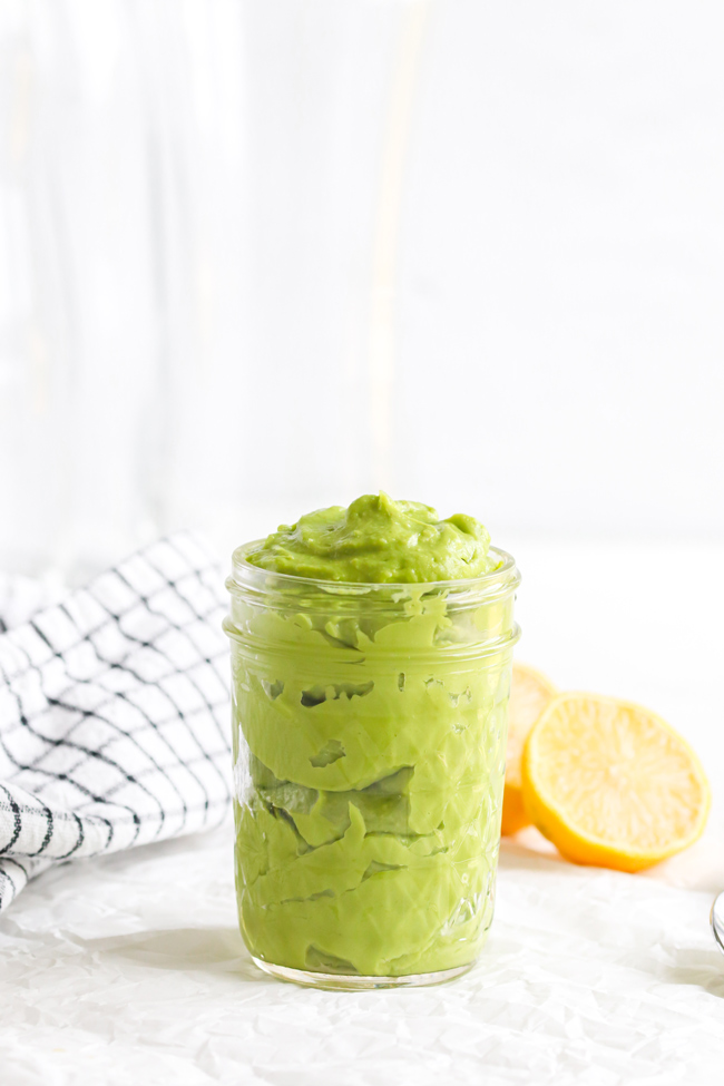 Avocado Mayo in a small glass jar next to fresh lemons and a black and white plaid cloth napkin.