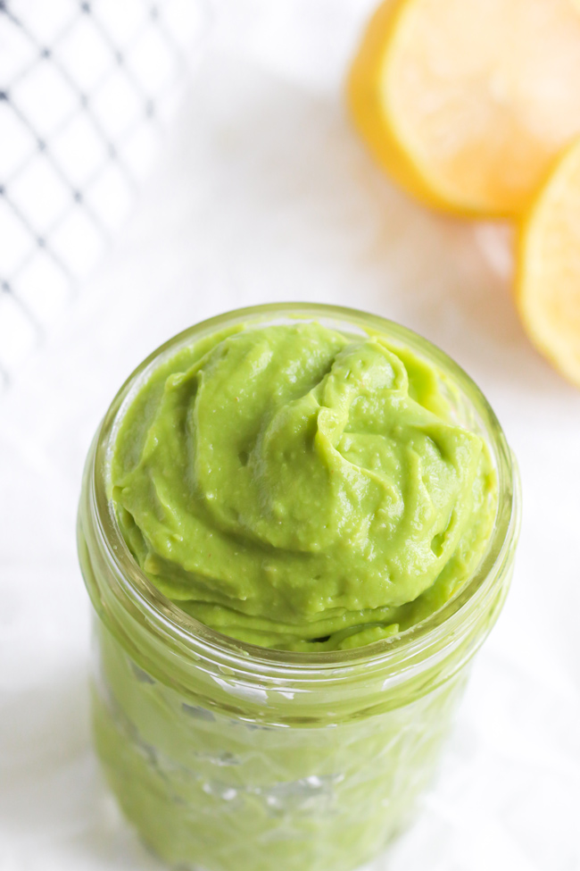 Avocado Mayo in a small glass jar next to fresh lemons and a black and white plaid cloth napkin.
