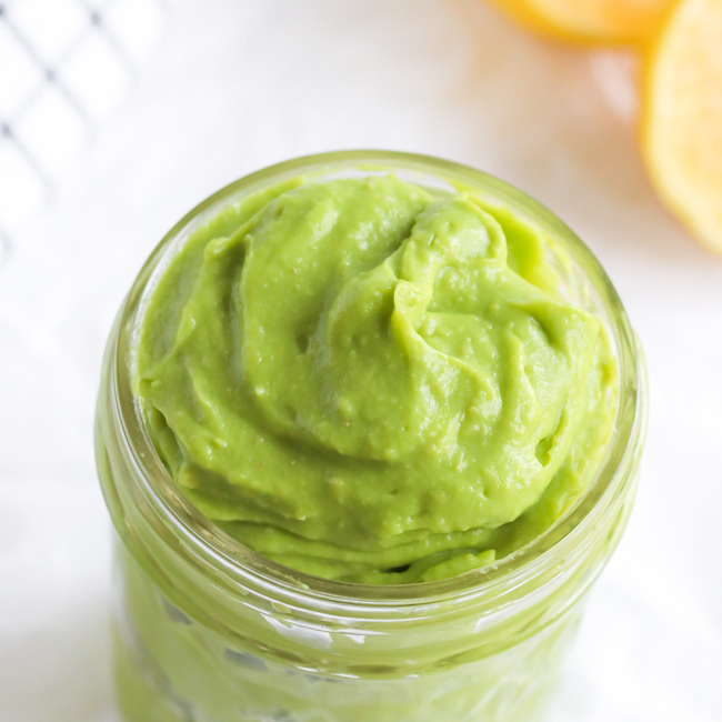 Avocado Mayo in a small glass jar next to fresh lemons and a black and white plaid cloth napkin.
