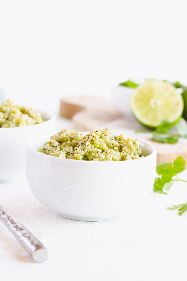 Creamy Avocado Cauliflower Rice in a small white bowl in front of a wooden cutting board, fresh limes, and cilantro.