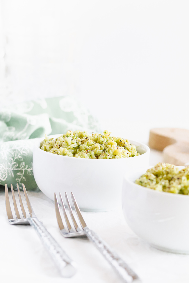 Creamy Avocado Cauliflower Rice in to small white bowls next to a green floral napkin and hammered silverware.