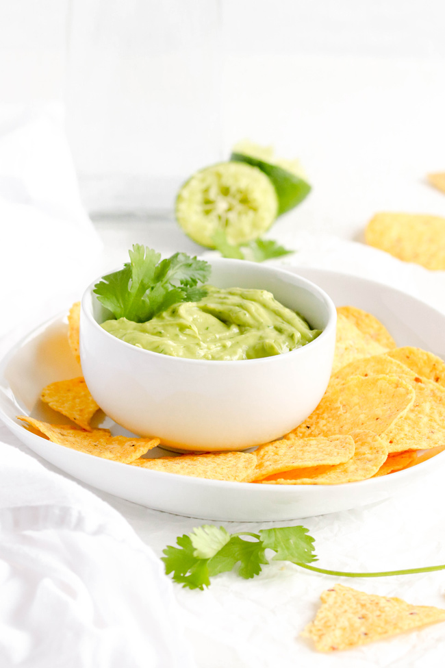 Creamy Avocado Lime Spread in a white bowl on a white plate surrounded by round yellow tortilla chips and a sprig of fresh cilantro.