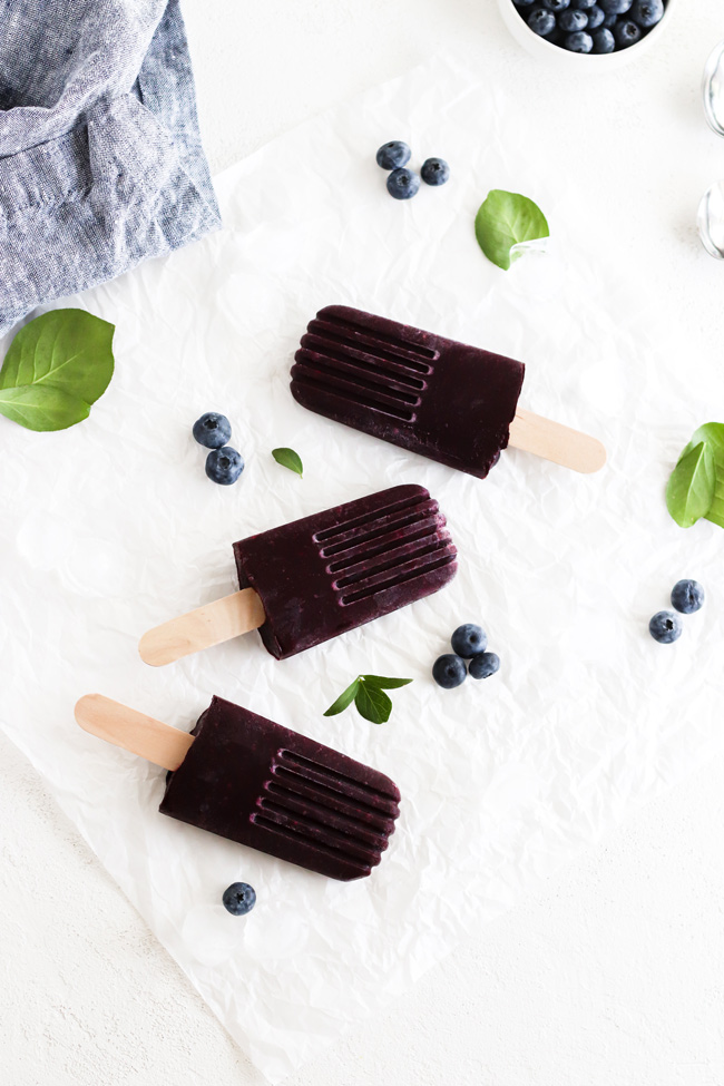 Dairy-Free Blueberry Popsicles on a piece of white parchment paper next to a cloth napkin, fresh blueberries, green leaves, and ice cubes.