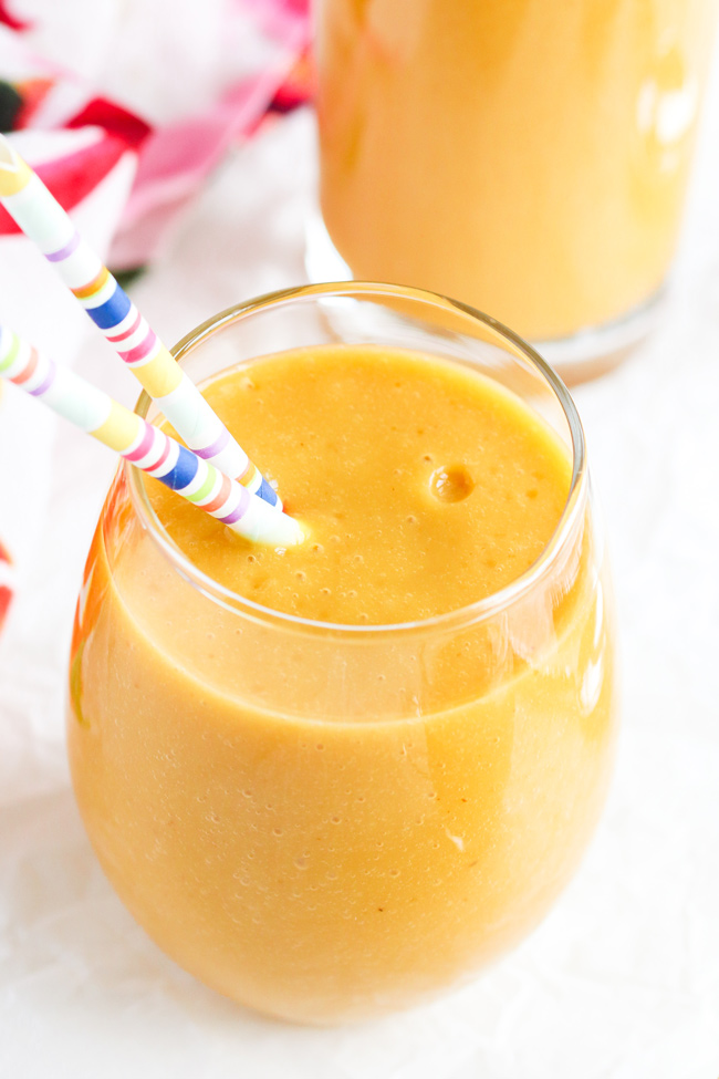 Peach Mango Smoothie in a drinking glass with striped paper straws, next to a floral napkin.