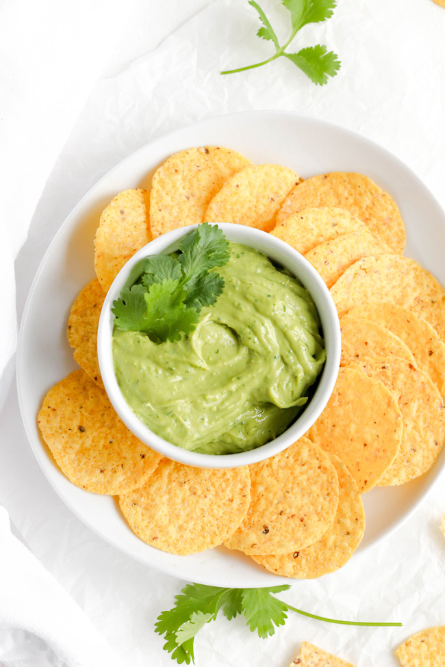 Creamy Avocado Lime Spread in a white bowl on a white plate surrounded by round yellow tortilla chips, a sprig of fresh cilantro, and a white dish towel.